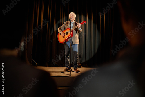Wide shot of Caucasian senior man performing standup music set, singing into microphone and playing acoustic guitar on stage. Useful for live entertainment, talent show, event promotion
