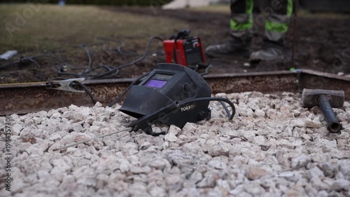 TORROS helmet rests on pale gravel by a rusted rail as leads run to a red unit, a sledgehammer lies nearby, and a worker in high vis trousers stands. Shallow depth of field.