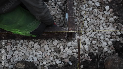 Overhead view shows a worker kneeling and adjusting yellow string lines on a rusted metal frame over crushed stone, with a clamp and welding cable visible in low light.