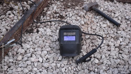 Close up shows worn welding helmet with blue tinted lens on pale crushed stone, rusted steel beam, sledgehammer, and welding lead, in neutral daylight.