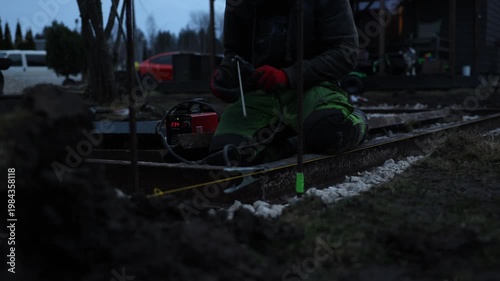 Worker kneels with helmet and red gloves, operates small red welder showing 234, guides rod on rusty rails, yellow string and green taped stake mark alignment.