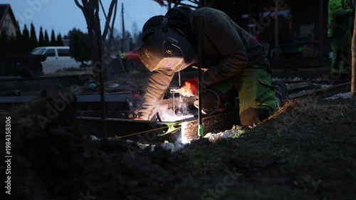 Worker kneels on rail track performing electric welding at night. Arc flare sends sparks and thin smoke as a measuring line holds alignment. Low angle, cool tones.