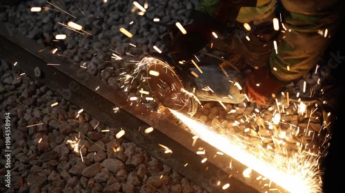 Close up shows worker grinding rail as sparks spray across crushed ballast. Makita logo is visible, heavy gloves protect hands, fast shutter freezes molten trails.