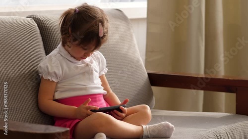 Three year old girl sits on a sofa watching cartoons on her father smartphone with full attention. The scene shows childhood, technology, and modern family life.