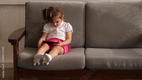 Three year old girl relaxes on a sofa watching cartoons on her mother smartphone. The scene shows comfort, childhood, and modern digital habits.