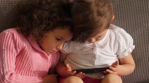 Two little sisters sit together watching cartoons on a smartphone. The scene shows modern childhood, family bonding, and shared digital entertainment.