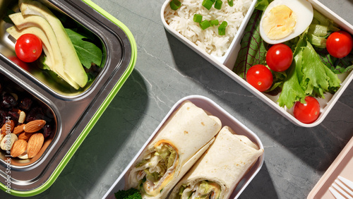 Healthy lunch boxes with vegetables, rice, and wraps on a gray surface