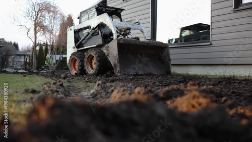 Low angle view shows skid steer with mud filled bucket moving along house siding near greenhouse, fresh furrows and deep tire tracks in late afternoon light.