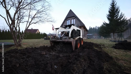 Skid steer with rusted bucket pushes churned soil near steep roof house. Operator sits in cab. Leafless tree left, tall conifer right, wide angle, cold light.