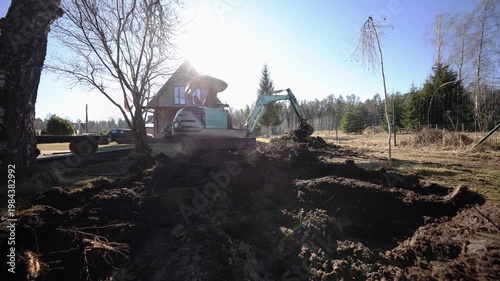 Small turquoise excavator digs soil by triangular roof wooden house. Low angle wide frame shows root filled earth, birch trunks, wire fence, lens flare, and active arm.
