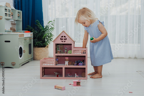 Child girl in blue dress arranges wooden furniture in a doll house.