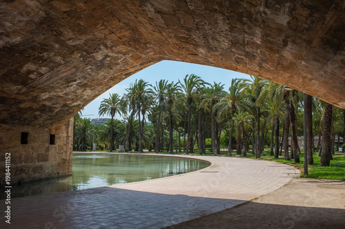 View of a pond and palm trees under a stone bridge arch in Turia Garden. Scenic public park landscape on a sunny summer day. Valencia. Spain. Europe.