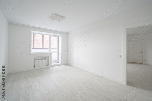 A bright, empty room with white walls and ceiling, light wooden flooring, a large window with a radiator, a ceiling light fixture, and an open doorway leading to another room