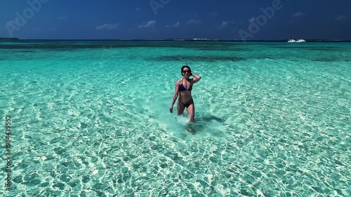 Woman walking through clear shallow turquoise ocean water on Maldives beach under sunny sky. Concept of travel, vacation, summer, tourism, relaxation, tropical lifestyle.