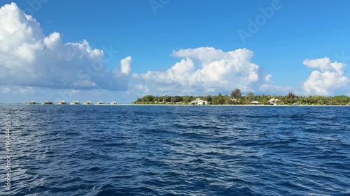 View from moving boat over ocean toward tropical Maldives island with villas and blue sky. Concept of travel, tourism, vacation, seascape, island life, journey.