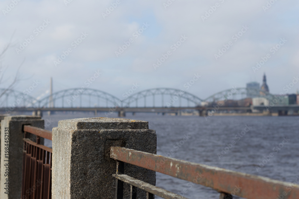Fototapeta premium Close-up of concrete embankment post and rusty railing beside river, with softly blurred arched bridge and skyline in background.