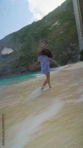 Young woman running on the beach below Kelingking cliffs on Nusa Penida, Bali, enjoying ocean waves and tropical freedom.
