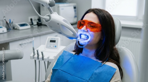 Woman undergoing professional teeth whitening procedure in dental clinic. Patient with protective goggles and cheek retractor using UV light technology for bright smile.