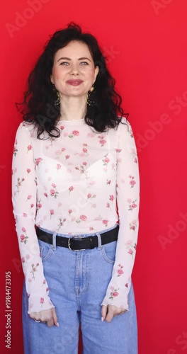Playful young woman with curly black hair winking and smiling at camera on red background. Beautiful girl in floral sheer blouse and blue jeans posing with cheerful facial expressions in studio.