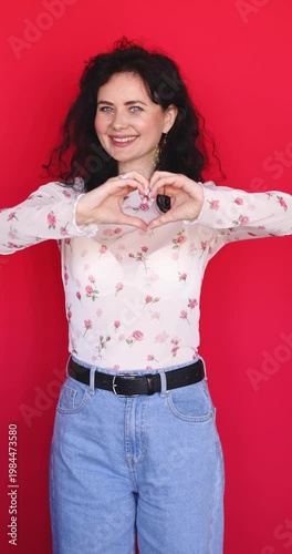Cheerful young woman showing heart shape with hands and smiling happily on red background. Beautiful brunette girl in floral sheer blouse expressing love, kindness and positive emotions in studio