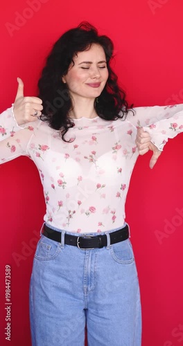 Pretty young woman showing thumb up and thumb down at same time on red background. Brunette girl in floral sheer blouse balancing between good and bad choice or feedback, comparing options indoor