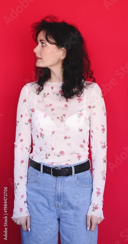Young woman in rose print mesh top looking left and right with curious gaze then showing shocked face expression on red background. Curly hair model in jeans posing for camera in studio.