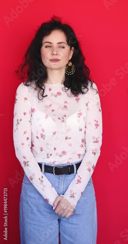 Pretty young woman with curly hair nodding head in agreement and smiling cheerfully on red background. Beautiful girl in floral sheer top giving yes gesture and looking at camera.
