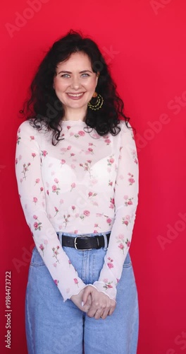 Cheerful woman with curly hair laughing and posing playfully on red background. Happy young female model in floral mesh top and jeans smiling with joy, showing sincere positive emotions in studio.