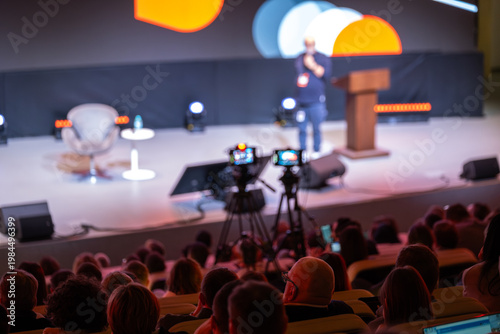 Photos Conference speaker on stage, cameras recording presentation, live event atmosphere, attentive audience, modern lighting and audiovisual production