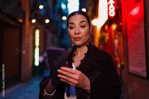 Obraz na plátně Hispanic woman with intrigued expression reading from tablet on street at dusk, symbolizing discovery, tech fluency and curiosity in modern connected lifestyle