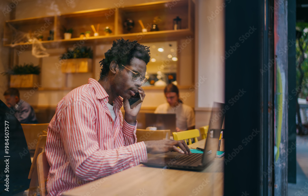 Fototapeta premium Young man on smartphone at laptop, multitasking in café setting. Technology‑enabled communication, remote work connection and mobile productivity in a casual workspace.