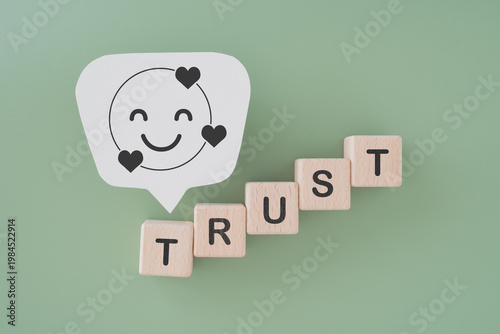 TRUST text on wooden cube block and smile face icon with heart on real white speech bubble paper with green background for customer satisfaction, service, feedback concept