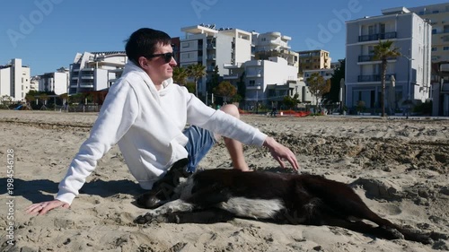 Happy young man petting stray dog on sunny beach.