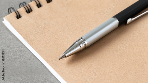 A silver black metal pen rests diagonally on a tan spiral notebook, ready to write.