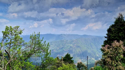A wide scenic view in Mukteshwar, Uttarakhand captures layered green mountain ridges stretching into distance beneath a sky filled with soft, billowing clouds, framed by tall trees, hillside foliage.