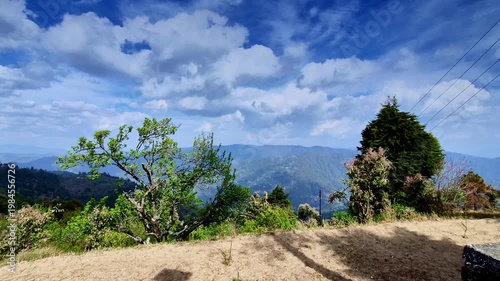 A breathtaking panoramic view in Mukteshwar, Uttarakhand showcases rolling green mountain ranges under a dramatic sky filled with fluffy clouds, with scattered trees and dry grassy foreground.