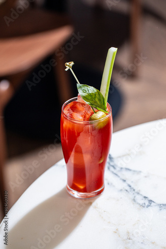 Red bloody mary cocktail with celery olive and ice served in tall glass on table in bar interior
