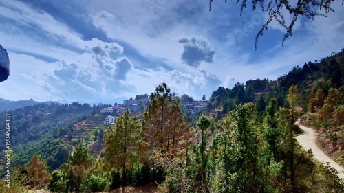 A sweeping hillside view in Mukteshwar, Uttarakhand shows terraced fields, scattered houses, and pine-covered slopes under a dramatic sky filled with layered clouds, with a winding dirt path.