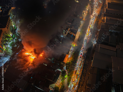 Top-down aerial angle capturing fire scene in Hanoi, flames spreading across rooftops while dense traffic flows along adjacent illuminated roadway.