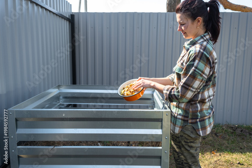 Adding kitchen food scraps and grass to metal compost bin, home composting process, sustainable organic waste reuse in steel frame structure.