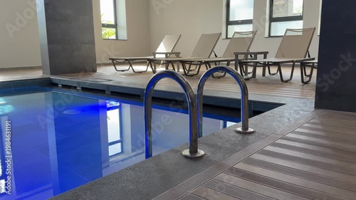 Indoor swimming pool with metal handrails, blue water, and lounge chairs along wooden deck