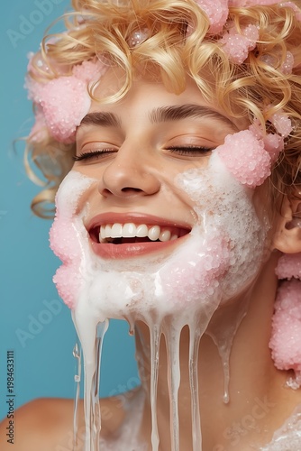 Smiling woman with curly blonde hair and pink face mask dripping with white cream on blue background