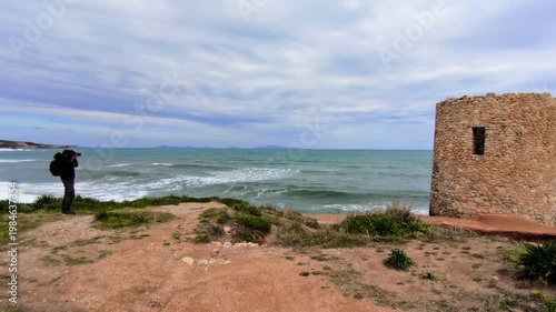 Rear view of a photographer in weather resistant gear standing on a rocky shore, capturing the power of a storm with crashing waves and a dark, turbulent sky over the sea