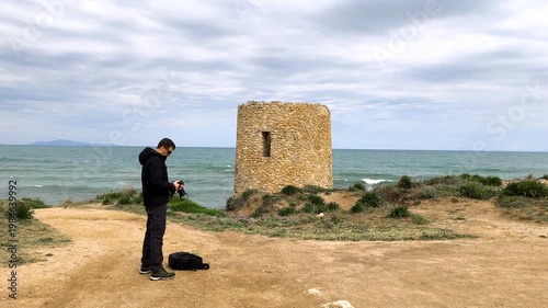 Rear view of a photographer in slow motion on a coastal dirt path, checking his camera under a dramatic, cloudy sky with the sea in the background.