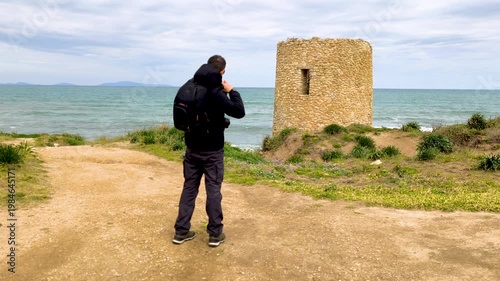 Rear view of a photographer putting on his backpack in slow motion on a coastal path, with the Mediterranean sea in the background on a sunny day.