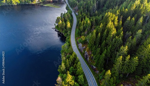 Aerial drone view of asphalt road winding alongside serene lake and dense evergreen forest during golden hour sunset light