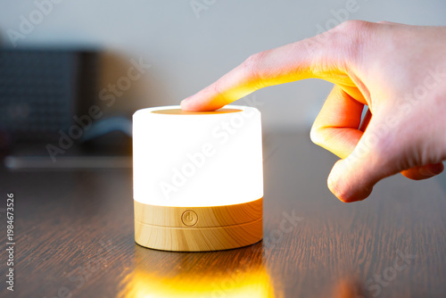 Bild auf Leinwand Hand touching the top of a glowing cylindrical wooden touch lamp on a dark table
