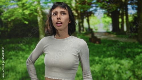 Fotografi Woman in gray long sleeve top raises hands in green forest clearing under bright sunlight; uncertainty doubt confusion hesitation