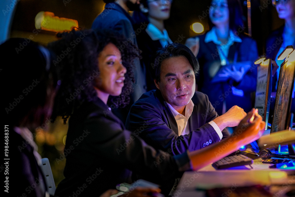Fototapeta premium Dedicated male and female professionals discussing project strategy at night. Crowded IT agency workplace with diverse employees analyzing data on glowing screens.