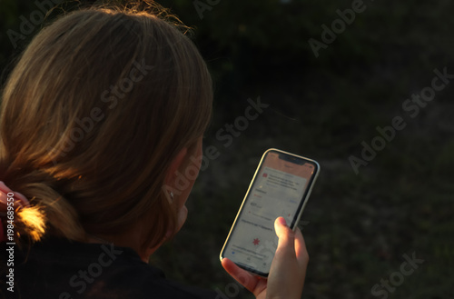 Over-the-shoulder view of a teenage girl using a smartphone outdoors at sunset. Teen looking at screen, scrolling social media feeds. Digital addiction, communication